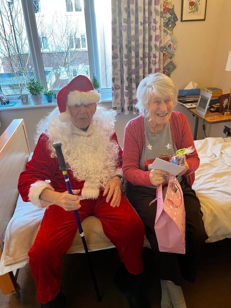 santa sat down on a female residents' bed smiling with her after delivering a Christmas gift