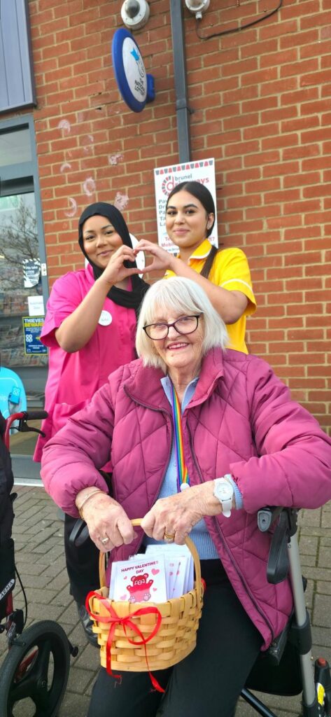woman wearing a pink coat out in the community handing out homemade biscuits with 2 team members making a heart with their hands behind her
