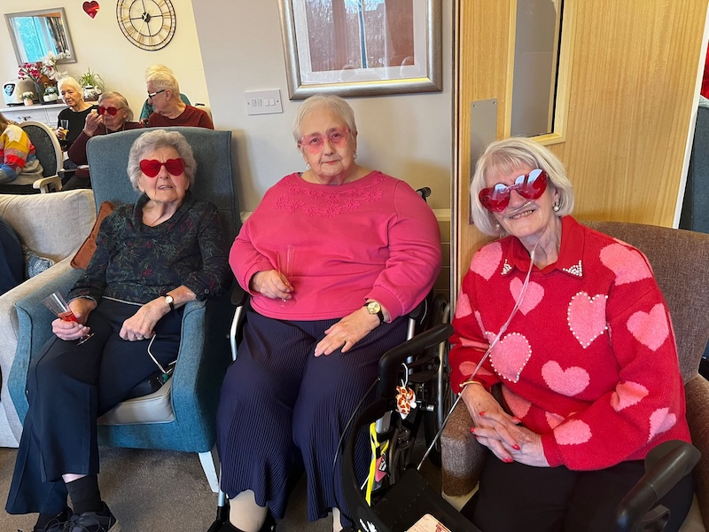 3 of our residents sat in armchairs in our lounge in valentines themed outfits and heart glasses smiling