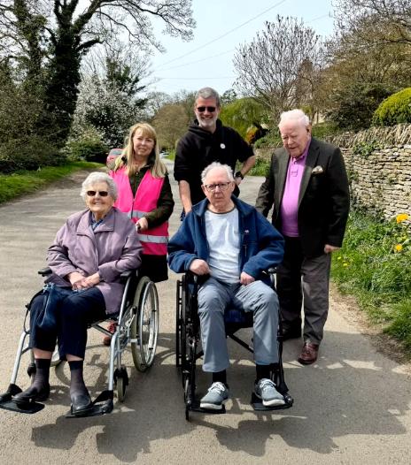 two older adults in wheelchairs and three others stood with them on a path